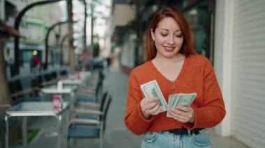 Young redhead woman smiling confident counting dollars at street
