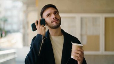 Young hispanic man drinking coffee listening audio message by the smartphone at street