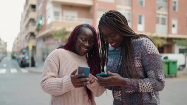 Two african american women smiling confident using smartphone at street