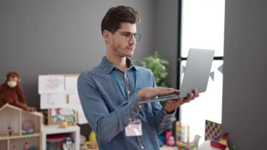Young hispanic man preschool teacher using laptop standing at kindergarten