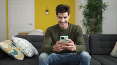 Young hispanic man using smartphone doing ok gesture at home