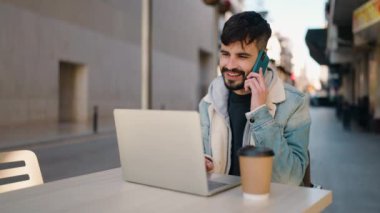 Genç İspanyol adam dizüstü bilgisayar kullanıyor ve kahve dükkanının terasında telefonla konuşuyor.