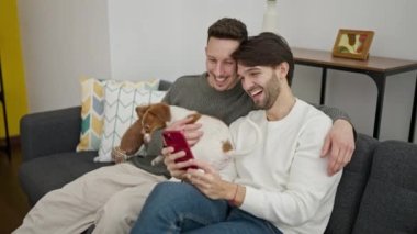İki erkek akıllı telefon kullanıyor. Kanepede köpeğiyle oturuyor.