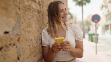 Beyaz bir kadın sokakta akıllı telefon kullanarak gülümsüyor.