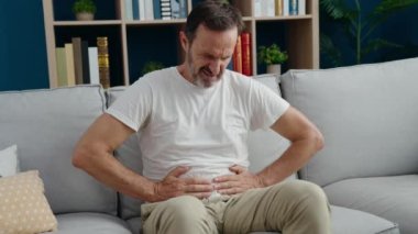 Orta yaşlı bir adam mide ağrısı çekiyor. Evdeki kanepede oturuyor.