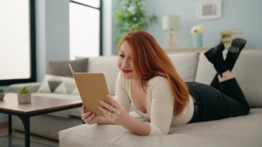 Young redhead woman reading book lying on sofa at home