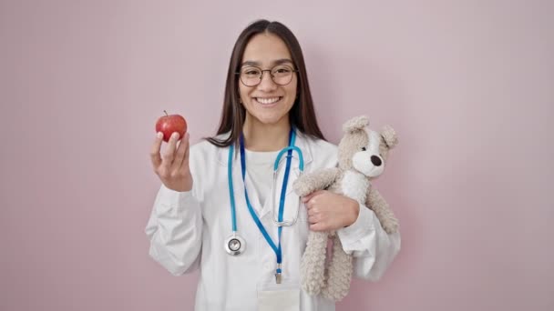 年轻美丽的惊慌失措的女医生微笑着 自信地抱着泰迪熊和苹果 披着孤立的粉色背景 — 图库视频影像