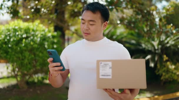 若い中国人男性宅配便使用スマートフォン保持パッケージで公園 — ストック動画