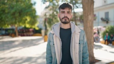 Genç İspanyol adam gülümsüyor. Parkta kollarını kavuşturmuş duruyor.