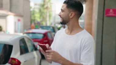 Genç Arap adam sokakta akıllı telefon kullanarak kendinden emin gülümsüyor.