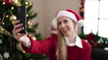 Young blonde woman having video call sitting on sofa by christmas tree at home