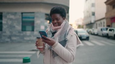 Genç Afrikalı Amerikalı kadın akıllı telefon kullanıyor ve sokakta kahve içiyor.