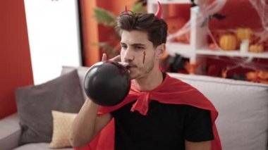 Young hispanic man wearing devil costume inflating balloon at home