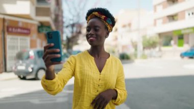 Genç Afrikalı Amerikalı kadın gülümsüyor. Kendine güveni tam. Sokakta akıllı telefondan selfie çekiyor.