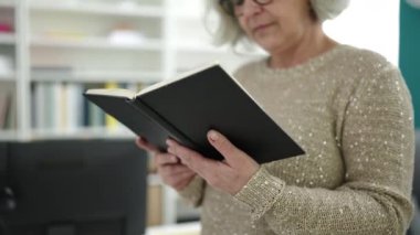 Middle age woman with grey hair teacher reading book doing silence gesture at university classroom