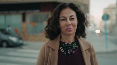 Orta yaşlı İspanyol kadın gülümsüyor. Kollarını kavuşturup caddede duruyor.