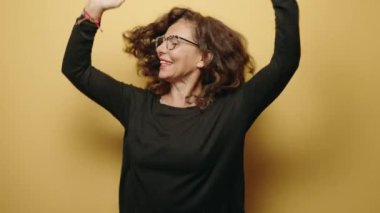 Orta yaşlı kadın gülümsüyor, sarı arka planda dans ediyor.