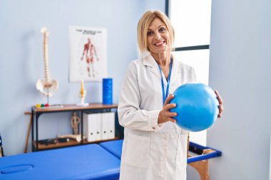 Middle age blonde woman wearing physiotherapist uniform holding ball at physiotherapy clinic