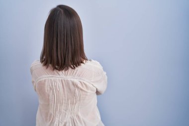 Orta yaşlı Latin bir kadın mavi arka planda dikilip kollarını kavuşturup arkasını çeviriyor. 