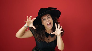 Middle age hispanic woman wearing witch costume doing scare gesture with hands over isolated red background