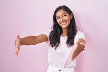 Pembe arka planda duran genç İspanyol kadın kameraya sarılmak için kollarını açıp gülümseyerek bakıyor. Mutluluğu kucaklayan neşeli bir ifade.. 