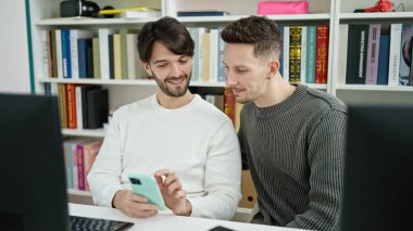 Kütüphanede bilgisayar ve akıllı telefon kullanan iki erkek öğrenci.