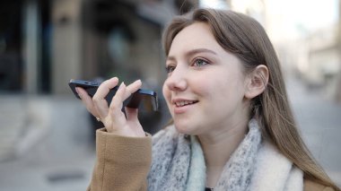 Young blonde woman smiling confident sending voice message by smartphone at street