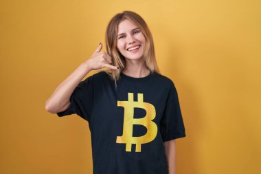 Sarışın, beyaz kadın bitcoin tişörtü giyiyor, gülümsüyor, telefonla el ve parmak hareketleri yapıyor, telefonda konuşuyor gibi. iletişim kavramları. 
