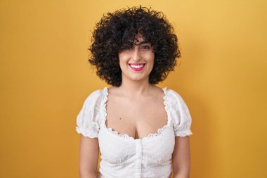Sarı arka planda kıvırcık saçlı, mutlu ve havalı bir gülümsemesi olan esmer bir kadın. şanslı kişi. 