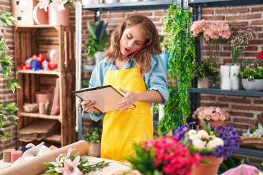Young beautiful hispanic woman florist talking on smartphone reading clipboard at florist
