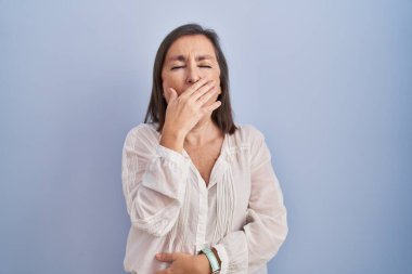 Mavi arka planda dikilen orta yaşlı İspanyol kadın ağzıyla ağzını kapatmaktan sıkılmış. Huzursuz ve uykulu.. 