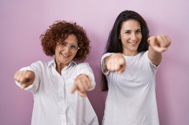 Hispanic mother and daughter wearing casual white t shirt over pink background pointing to you and the camera with fingers, smiling positive and cheerful 
