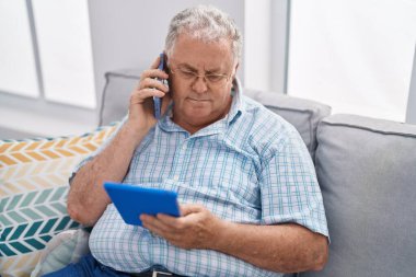 Middle age grey-haired man talking on smartphone using touchpad at home