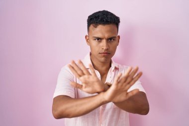 Young hispanic man standing over pink background rejection expression crossing arms and palms doing negative sign, angry face 