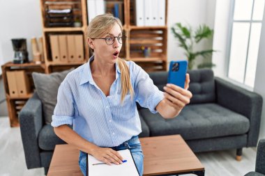 Muayenehanede online terapi seansı yapan güzel bir kadın ağzı açık kalmış, korkmuş ve şaşırmış bir şekilde şaşkınlık içinde şaşkınlık içinde. 