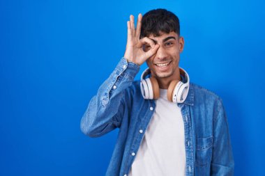 Young hispanic man standing over blue background doing ok gesture with hand smiling, eye looking through fingers with happy face. 