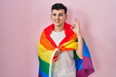 İkili olmayan kişi elinde gökkuşağı lgbtq bayrağı tutuyor tamam işareti yapıyor parmaklarıyla, gülümseyerek mükemmel bir sembol oluşturuyor 