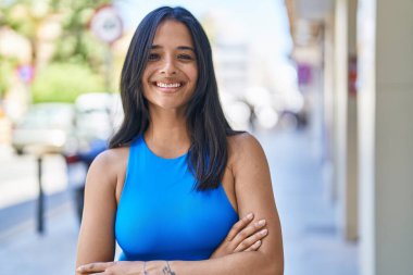 Genç İspanyol kadın gülümsüyor. Kollarını kavuşturup caddede duruyor.