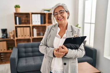 Muayenehanede gri saçlı orta yaşlı bir kadın kameraya seksi bir ifadeyle bakıyor, neşeli ve mutlu bir yüzle.. 