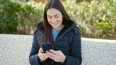 Genç beyaz kadın parkta akıllı telefon kullanarak gülümsüyor.