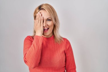 Sarışın kadın izole edilmiş arka planda duruyor. Bir gözünü eliyle kapatıyor. Yüzünde kendine güvenen bir gülümseme ve sürpriz bir duygu var.. 