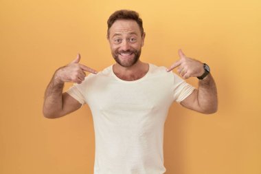 Sarı arka planda sakallı orta yaşlı bir adam. Yüzünde bir gülümsemeyle gururlu ve mutlu birini işaret ediyor.. 