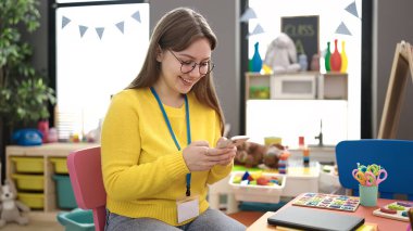 Young blonde woman preschool teacher smiling confident using smartphone at kindergarten