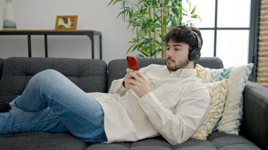 Genç İspanyol adam evde kanepede uzanmış akıllı telefonuyla müzik dinliyor.