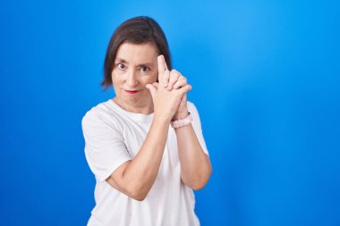 Middle age hispanic woman standing over blue background holding symbolic gun with hand gesture, playing killing shooting weapons, angry face 