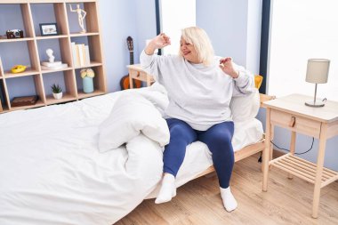 Middle age blonde woman waking up stretching arms at bedroom