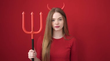Young blonde woman wearing devil costume holding trident over isolated red background