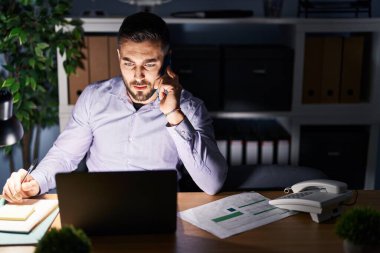 Young hispanic man business worker talking on the smartphone working overtime at office
