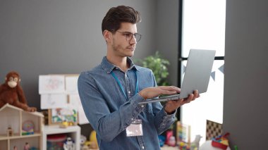 Young hispanic man preschool teacher using laptop standing at kindergarten