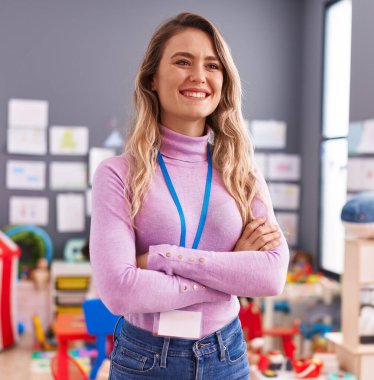 Young blonde woman teacher smiling confident standing with arms crossed gesture at kindergarten